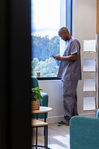 African american healthcare worker using smartphone by window in hospital lounge