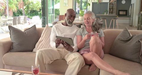Diverse senior couple relaxing on sofa, sipping pink drinks and browsing tablet in open-plan living