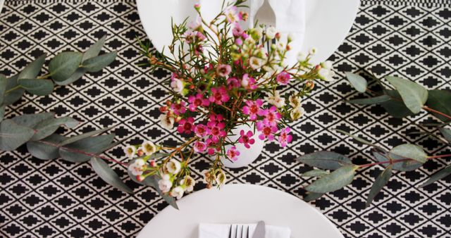 A beautifully arranged table setting features a floral centerpiece with vibrant pink flowers and lush greenery, with copy space. The geometric patterned tablecloth adds a modern touch to the elegant dining presentation.