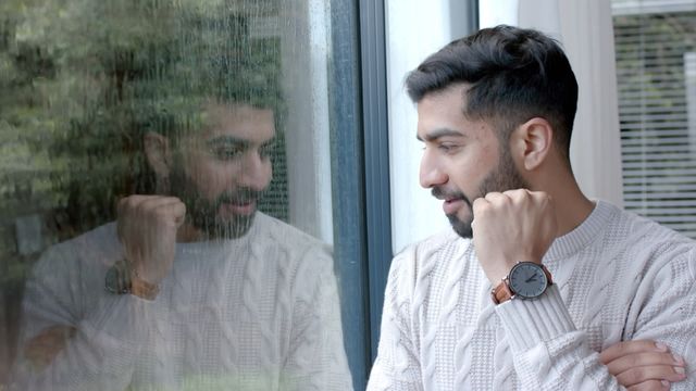 Man standing by window engaged in deep thought. Wearing casual knit sweater with wristwatch serving as sole accessory. Reflection creating introspective impression that underscores mood, ideal for lifestyle blogs, mental health discussions, or domestic life features.