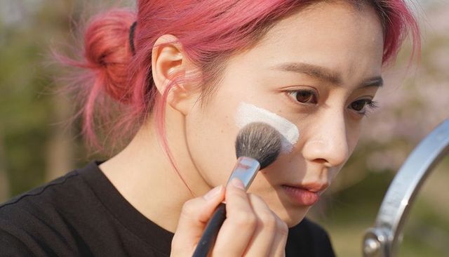 Young woman applying sunscreen with makeup brush outdoors, blending cream on cheek