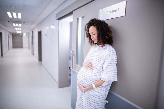 A pregnant woman in a hospital gown is standing in a corridor, waiting. Ideal for use in healthcare, maternity, and medical contexts to convey themes of pregnancy, patient care, and hospital environments.