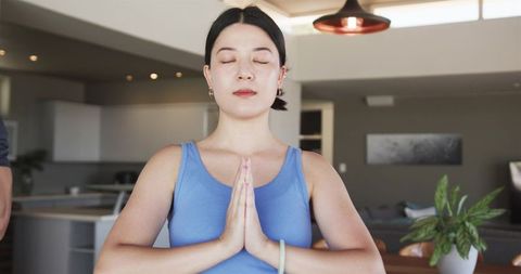 Asian woman meditating at home with hands in prayer pose in modern minimalist interior