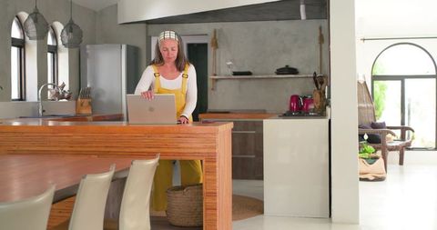 Woman working remotely at kitchen island in yellow overalls, modern rustic open-plan interior