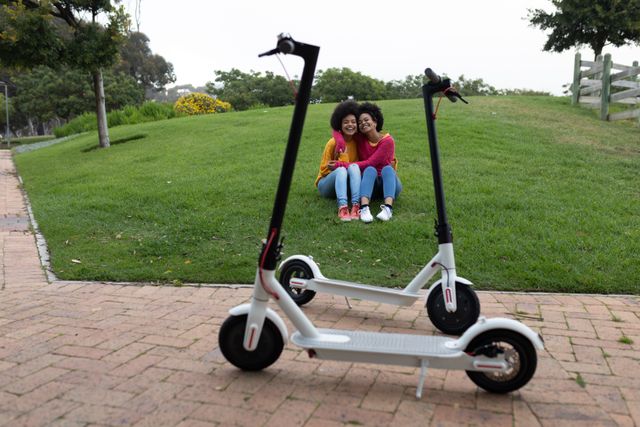 Front view of two happy biracial twin sisters enjoying free time together, embracing, smiling, sitting on grass in an urban park, electric scooters on a path in the foreground.
