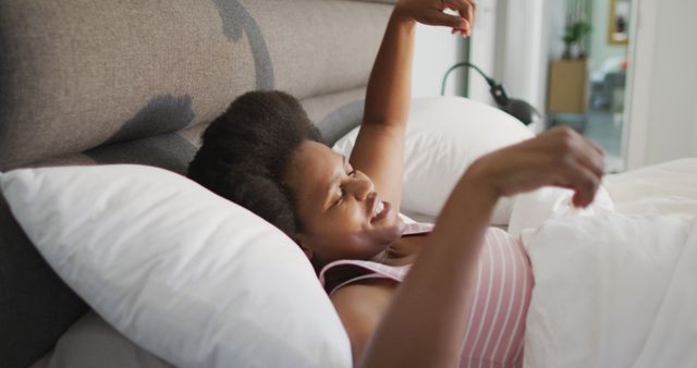 An African American woman is lying in bed and stretching her arms. She looks relaxed and is wearing casual clothes. The bedroom is simple and light, creating a peaceful and comfortable atmosphere. This image is perfect for promoting healthy morning routines, relaxation products, bedroom decor, or lifestyle content.