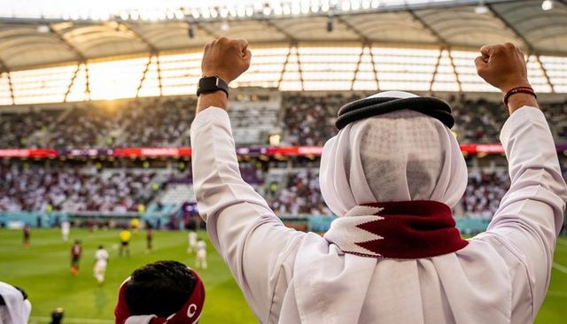 Cheering qatari football supporter wearing thobe and maroon scarf raising fists in stadium