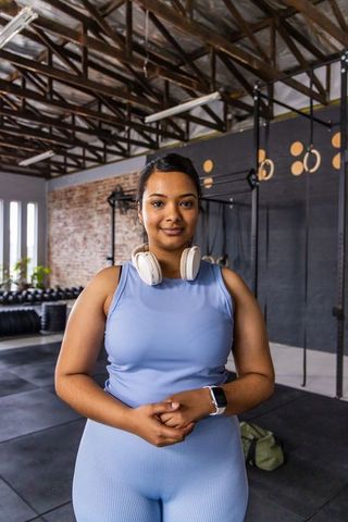 Confident asian woman in light blue activewear posing in gym with headphones and smartwatch