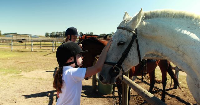 Two children wearing helmets patting and bonding with horses in an outdoor ranch environment. Sun is shining, setting an outdoor scene filled with equestrian themes, suitable for topics on farm life, children learning about animals, and benefits of equestrian activities.