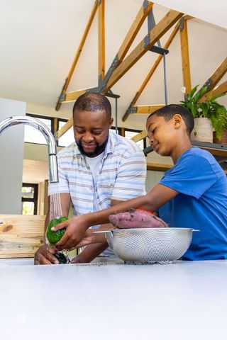 African american father and son rinsing vegetables at kitchen sink, bonding while cooking