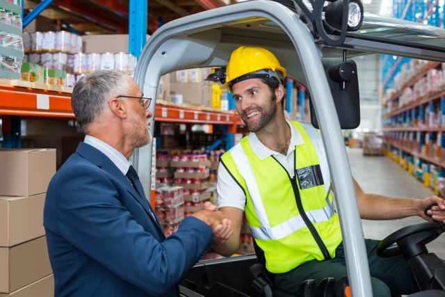 Warehouse worker in safety vest and hard hat shaking hands with manager in suit, symbolizing teamwork and collaboration in logistics and supply chain management. Ideal for use in articles or presentations about business partnerships, industrial operations, and workplace teamwork.