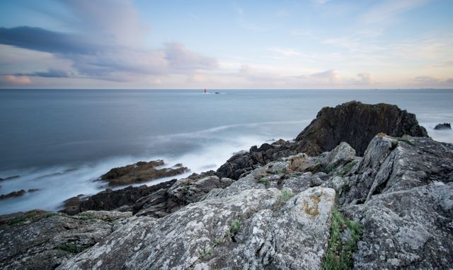 Rocky coastline during dawn with calm ocean and cloudy sky. Rugged rocks with gentle waves crashing, and horizon lit with soft morning light. Perfect for travel advertisements, nature posters, landscape paintings, meditation apps, and environmental awareness campaigns.