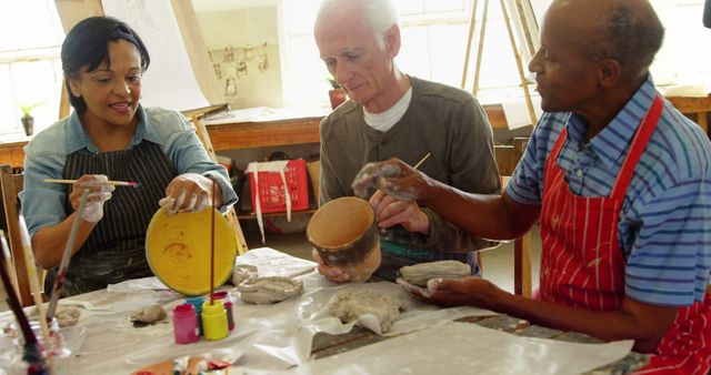 Group of senior citizens participating in a pottery class, painting and molding clay objects. Mixture of materials including brushes, paint, and clay on table. Bright and engaging atmosphere captures sense of community and creativity. Ideal for promoting elder leisure activities, art education, group therapy, and community centers.