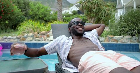 Relaxing african american man lounging by private pool in sunglasses and pink shorts