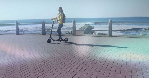 Woman riding e-scooter on seaside promenade in yellow jacket