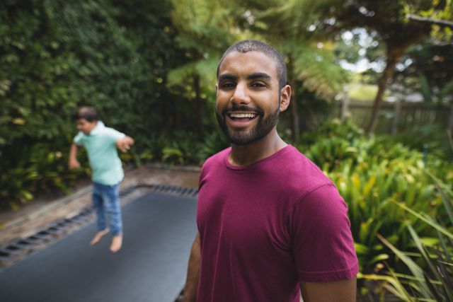 Father smiling while watching his son jump on a trampoline in a park. Ideal for use in family-oriented advertisements, parenting blogs, outdoor activity promotions, and lifestyle articles focusing on family bonding and outdoor fun.