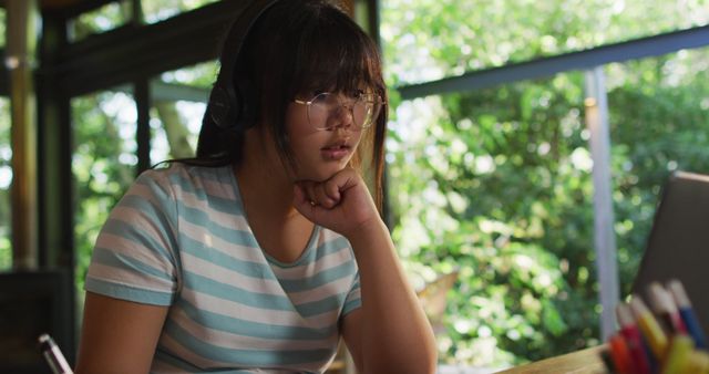 Young girl wearing glasses and headphones focuses on studying with laptop in bright room. Suitable for educational content, online learning platforms, technology in education themes, and teenage academic life scenes.