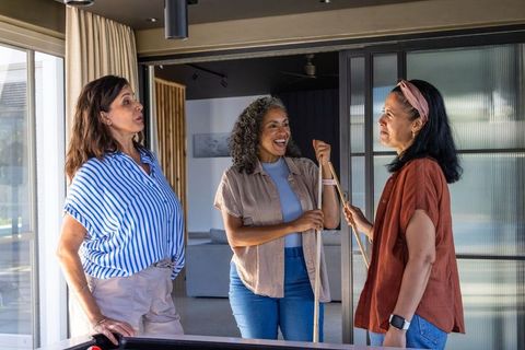 Multicultural women laughing and chatting around pool table in modern home game room