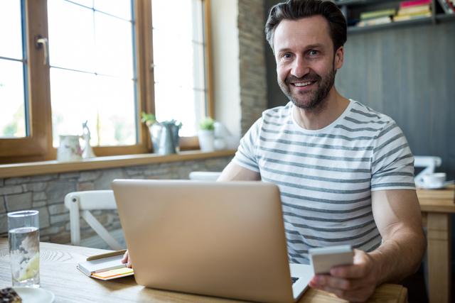 Man sitting at wooden table in coffee shop using laptop and holding mobile phone. Scene depicts modern remote work and digital nomad lifestyle. Ideal for use in articles about remote working, freelancing, technology in coffee shops, and modern workspaces.