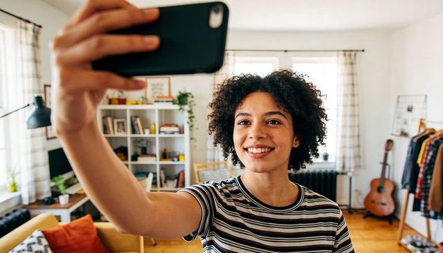 Young woman taking selfie in bright home studio with guitar, bookshelf and cozy decor
