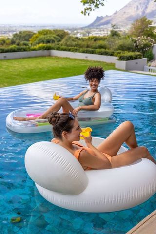 Diverse women relaxing on inflatable pool loungers with citrus drinks in backyard pool