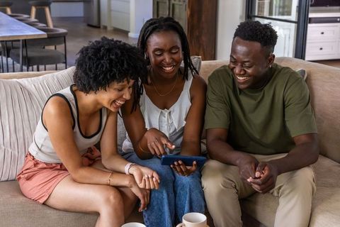 African american friends laughing together on sofa sharing smartphone and coffee