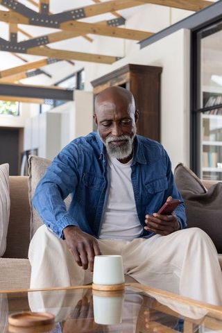 Senior african american man relaxing on sofa holding smartphone and ceramic mug