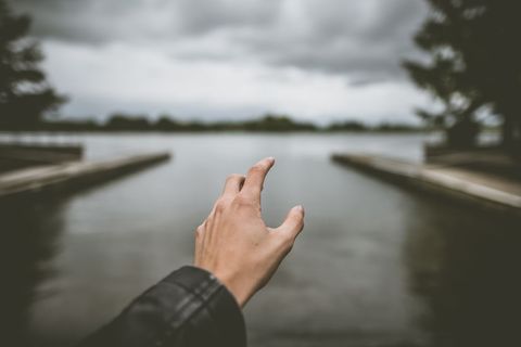 Outstretched hand reaching towards tranquil lake under cloudy sky