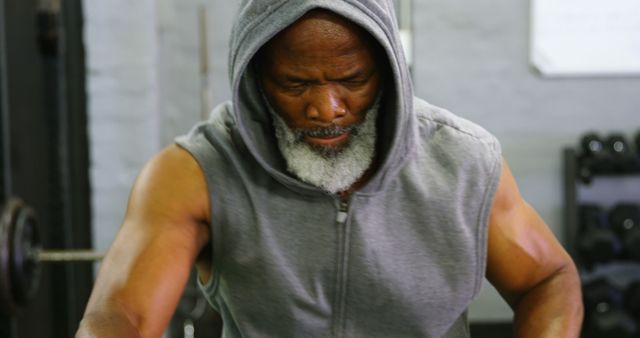 Senior man lifting weights at the gym, wearing a sleeveless hoodie. Ideal for use in fitness, health, and active lifestyle promotions targeting older adults. Useful in gym advertisements, workout apparel promotions, health and wellness articles, and motivational content for seniors.