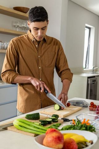 Young man slicing cucumber on kitchen island preparing fresh vegetables for healthy meal