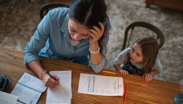 Stressed mom filling paperwork at kitchen table while young daughter watching and waiting