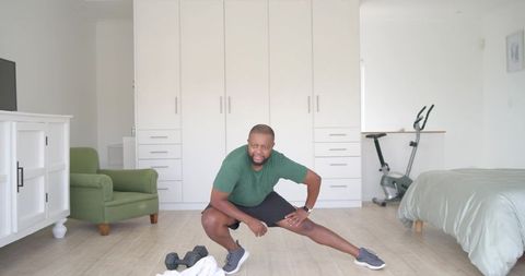 African american man stretching before home workout in bright bedroom with dumbbells and bike