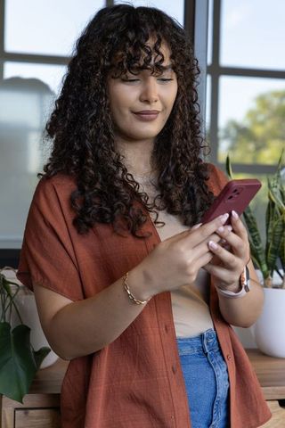 Curly-haired woman standing and texting on smartphone by sunny window with plants