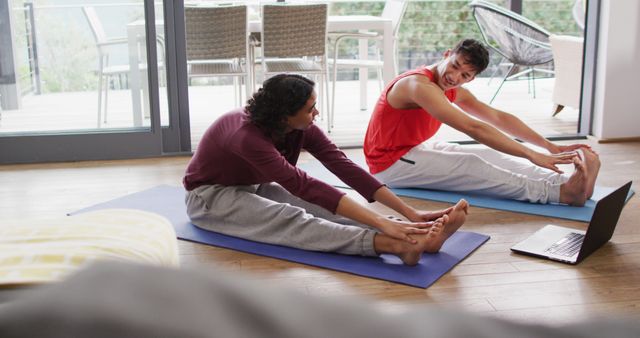 Friends are practicing yoga together at home on mats. They are stretching their legs and smiling at each other, creating a positive and relaxed atmosphere. A laptop is nearby, likely for virtual guidance or following an online workout. This image can be used to promote home fitness routines, yoga classes, virtual workout programs, healthy living, and social bonding activities.