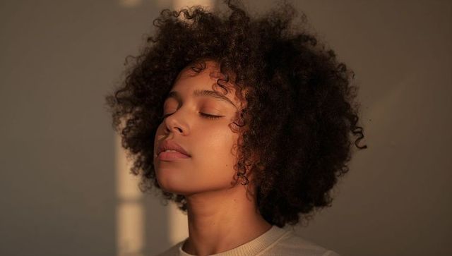 Serene young woman basking in golden hour light with curly hair, minimalist indoor portrait