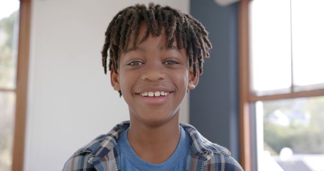 This photo shows a close-up of a smiling young boy with short dreadlocks. He's wearing casual clothes and standing indoors with natural light coming in from the windows behind him. Perfect for use in advertisements, websites, educational materials, or any project featuring joyful childhood moments and diversity.