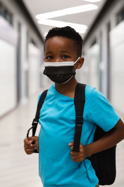 Young African American boy standing in a school hallway, wearing a protective mask and carrying a backpack. This image can be used for educational materials, back-to-school campaigns, health and safety guidelines, and articles related to childhood education during the pandemic.