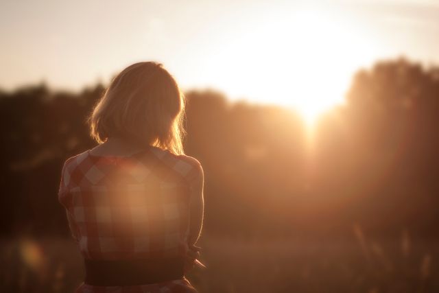 Young woman standing in field looking at sunset, sunlight creating lens flare. Ideal for illustrating concepts like tranquility, relaxation, inspiration, solitude, mental health, nature appreciation, and motivational content. Suitable for blogs, adverts, wellness articles, or nature-themed promotions.