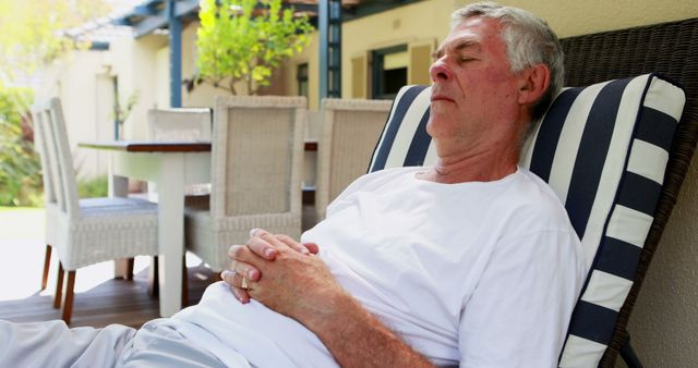 A senior Caucasian man is relaxing on a patio chair with his eyes closed, with copy space. His peaceful posture suggests a moment of rest or meditation in a tranquil outdoor setting.