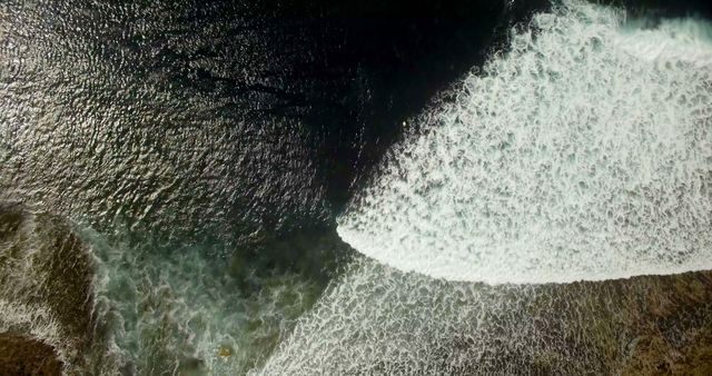Aerial capture of waves meeting the shore in dynamic motion. Ideal for projects focused on nature, coastline conservation, or travel promotions. The contrast between the dark ocean depths and white surf might be used to symbolize opposition or transition.
