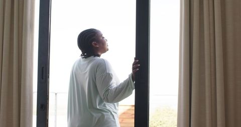 African american woman standing in doorway tilting toward sunlit balcony wearing loungewear