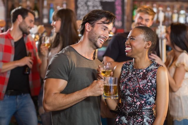 Young couple smiling and holding beer mugs while socializing with friends at a lively bar. Ideal for use in advertisements for bars, nightlife promotions, social events, and lifestyle blogs.