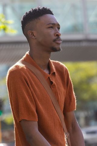 African man standing gazing right sunlit urban plaza wearing orange polo crossbody strap