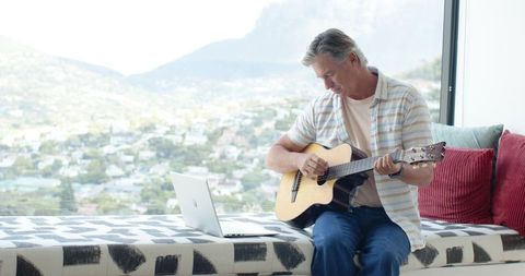 Mature man playing acoustic guitar using laptop for online lesson on sunlit window bench