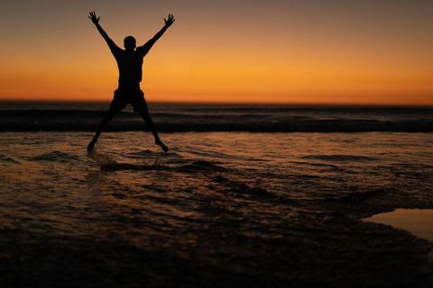 African American man jumping on beach at golden sunset silhouette splashing ocean waves