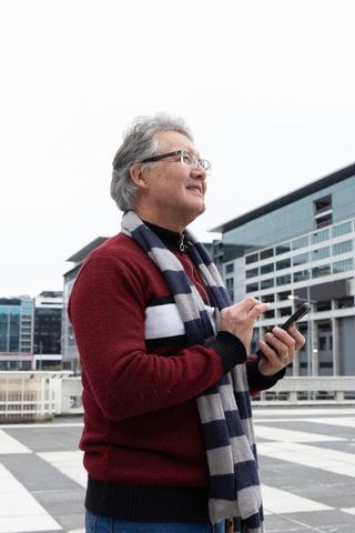 Senior asian man standing on urban rooftop using smartphone wearing red sweater and striped scarf