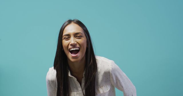 This image shows a young woman with long hair smiling and laughing joyfully against a vibrant turquoise background. She is dressed in casual attire, which adds to the relaxed and cheerful ambiance of the image. This photo is perfect for use in advertisements, social media posts, and marketing materials that aim to convey happiness, positivity, and a sense of carefree enjoyment. It can also be used in lifestyle blogs or personal growth articles to illustrate themes of joy and wellbeing.