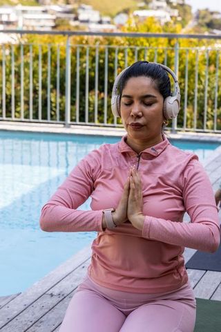 Meditating woman by pool wearing headphones and smartwatch in pink workout set