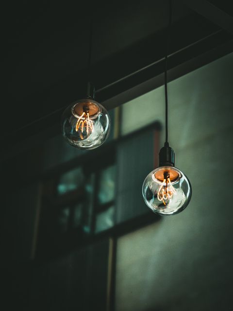 Two vintage Edison bulb hanging lights emitting warm light in a dark industrial space with an out-of-focus window backdrop. Suitable for illustrating interior design concepts, adding mood to marketing materials for cafés or bars, or showcasing retro and industrial decor themes in lifestyle magazines.