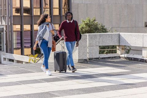 Diverse young friends walking through modern urban plaza pulling rolling suitcase and chatting
